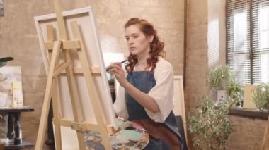 Orta uzunlukta kıvırcık kızıl saçlı beyaz kadın önlük giyiyor, gündüz vakti resim stüdyosunda fırçayla resim yapıyor.