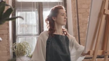 Önlük giyen kıvırcık saçlı genç bir kadının arka ışıklandırması, stüdyo tuvalinde macun bıçağıyla resim yapması.