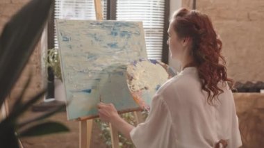 Önlük giyen kızıl saçlı, beyaz bir kadının, stüdyodaki tuvale macun bıçağıyla boya koyması.