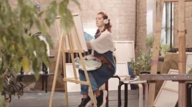 Kulaklık takan, müzik dinleyen, stüdyoda boya fırçasıyla resim yapan genç, kıvırcık, beyaz bir kadın.