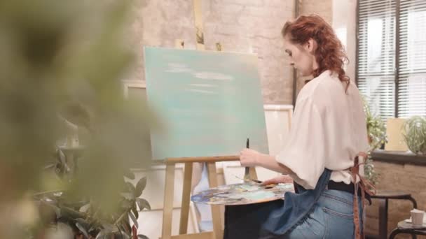 앞치마를 스튜디오에서 붓으로 미술을 앉아서 그림을 그리는 곱슬머리의 여성의 뒷모습 — 비디오