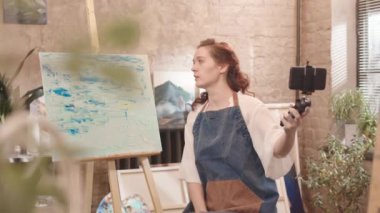 Beyaz önlük giyen, stüdyoda oturup resim yapan, konuşan, gülümseyen ve resimlerini akıllı telefon kamerasında gösteren beyaz bir kadın.