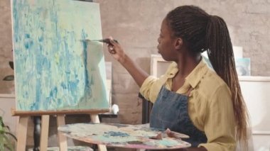 Önlük giyen, resim tekniği öğreten ve stüdyoda görünmez öğrencilerle konuşan genç Afrikalı Amerikalı kadın resim öğretmeninin izini sürmek.