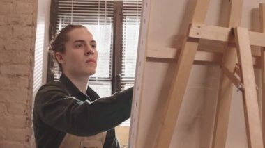 Gündüz vakti modern sanat stüdyosunun penceresinin önünde duran beyaz bir adam tuvalde oyuncak bıçakla resim yapıyor.