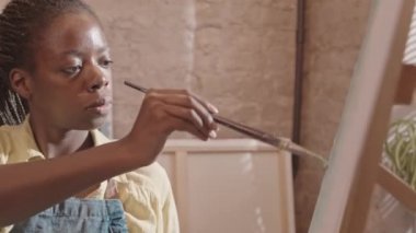 Gündüz vakti modern sanat stüdyosunda fırçayla resim yapan genç Afrikalı Amerikalı bir kadının orta boy yakın çekimi.