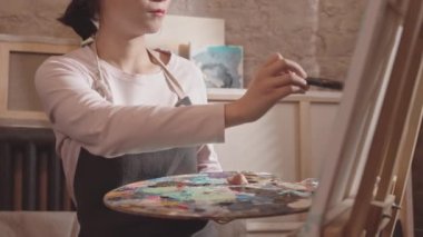 Modern sanat stüdyosunda önlük giyen, tuvalde fırçayla resim yapan genç Asyalı kadın sanatçıların orta boy yakın çekimi.