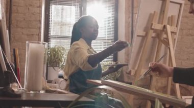 Düşük açılı genç Afrikalı Amerikalı kadın modern sanat stüdyosunda pencerenin önünde oturuyor, tuval üzerinde macun bıçağıyla resim yapıyor, ön planda kesilen öğrenci konuşuyor.