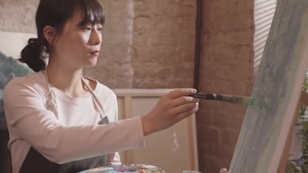 Střední detail mladé asijské ženy v moderním uměleckém studiu ve dne, malba štětcem na plátně