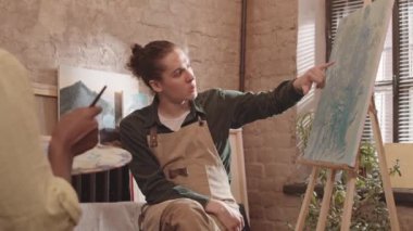 Orta boylu, kahverengi gözlü, önlük giyen, modern sanat stüdyosunda oturan, soyut tablosunu kırpılmış öğrencilere tarif eden beyaz bir adam.