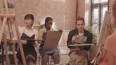 Orta boylu Asyalı bir kadın, Afro-Amerikalı bir kız öğrenci ve önlük giyen beyaz bir adam sanat sınıfında oturmuş, tanınmayan bir öğretmeni dinliyorlar.