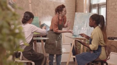 Orta boylu Asyalı ve Afrikalı Amerikalı genç sanat öğrencilerinin önlük giymesi, sınıfta oturması, sehpada tuvale resim çizmesi, kızıl saçlı beyaz bir akıl hocasının yürümesi ve onlara yardım etmesi.