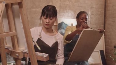 Resim dersinde resim yapan Asyalı genç bir kadın. Afrikalı Amerikalı arkadaşı resimlerini çekiyor. Önlük giyen öğrenciler.