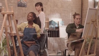 Orta uzunlukta beyaz erkek ve Afrikalı Amerikalı kadın önlük giyiyor, sınıfta oturuyor, sehpada tuvale resim çiziyor, Asyalı kadın akıl hocası yürüyor ve onlara tavsiyelerde bulunuyor.