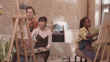 Orta uzunluktaki Asyalı ve Afrikalı Amerikalı genç sanat öğrencileri önlük giyiyor, sınıfta oturuyor, sehpada tuvale resim çiziyor, kızıl saçlı beyaz bir akıl hocası yürüyor ve onlara tavsiyelerde bulunuyor.