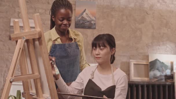 Közepes fehér férfi és ázsiai nő kötényben, osztályban ülve, festve vászonra festve az állványon, afro-amerikai női mentor sétál és segít nekik