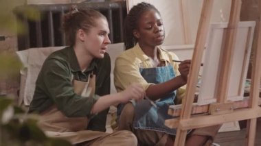 Önlük giyen Afrikalı Amerikalı kadın ressamın düşük açılı görüntüsü, tuvale boya fırçasıyla resim çizmesi, stüdyoda genç beyaz bir adamla oturması ve yardım etmesi.