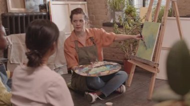 Kızıl saçlı beyaz kadın önlük giyiyor, resim stüdyosunda yerde oturuyor, boya ve fırçayla soyut resimler çiziyor, etnik çeşitliliğe sahip yetişkin öğrencilerle konuşuyor.