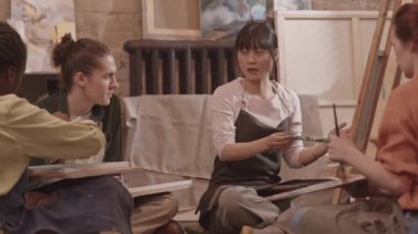 Önlük giyen, resim stüdyosunda yerde oturan, boya ve fırçayla soyut resim yapan, kesilmiş yetişkin öğrencilere ders veren ve onlarla konuşan mutlu Asyalı kadınların düşük açılı görüntüsü.