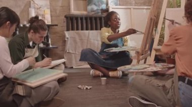 Önlük giyen, resim stüdyosunda yerde oturan, boya ve fırçayla resim yapan, etnik çeşitliliğe sahip yetişkin öğrencilerle konuşan Afrikalı Amerikalı kadınların düşük açılı görüntüsü.