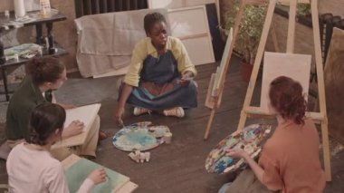 Önlük giyen, resim stüdyosunda yerde oturan, boya ve fırçayla soyut resim çizen, etnik çeşitliliğe sahip yetişkin öğrencilere öğreten ve onlarla konuşan Afrikalı Amerikalı kadınların yüksek açısı.