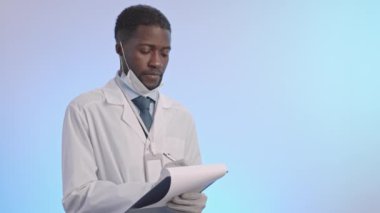 Tilt up medium slowmo portrait of smiling young African-American doctor in lab coat writing on clipboard then looking at camera standing on clear background