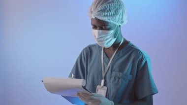 Tilt up medium portrait of young African-American male surgeon in uniform and face mask writing on clipboard standing on light blue background in studio