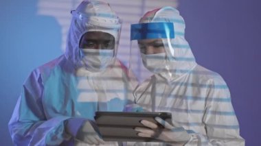 Medium shot of two young diverse medical workers in protective costumes, face shields and masks looking at digital tablet while having conversation indoors