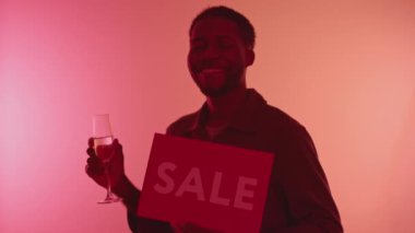 Medium slowmo portrait of cheerful African-American young man holding glass of champagne and red sale sign dancing at camera on neon pink background