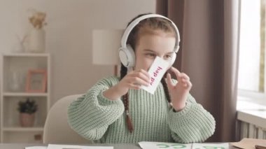 Waist-up slowmo shot of happy 5-year-old Caucasian girl in headphones having online speech therapy lesson on laptop, practicing pronunciation on words printed on cards
