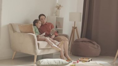Slowmo shot of 5-year-old girl and female speech therapist practicing correct pronunciation of sounds using mirror, sitting together on sofa in cozy living room