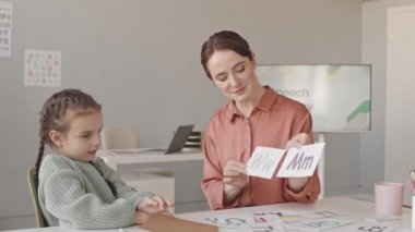 Waist up slowmo shot of little girl studying alphabet with female speech therapist having lesson in classroom