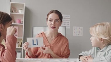 Waist up slowmo shot of two cute little boys studying correct articulation and pronunciation of letters with young female speech therapist in classroom