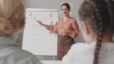 Medium slowmo shot of young female speech therapist giving lesson to children in classroom, standing by whiteboard with letters and sounds written on it