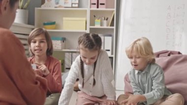 Medium slowmo shot of group of elementary kids and female speech therapist playing educational game using printed cards with words, sitting together on carpet in cozy classroom