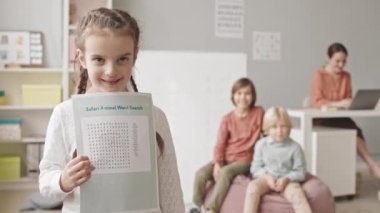 Medium slowmo portrait of 5-year-old girl posing for camera with word search worksheet standing in classroom of speech therapy with two boys and teacher in background