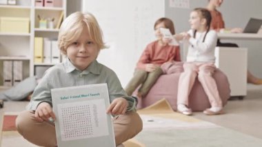 Slowmo portrait of 7-year-old Caucasian boy posing for camera with word search worksheet in hands having lesson of speech therapy with other kids and female teacher