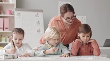 Waist up slowmo shot of three elementary age kids doing exercises on speech therapy in classroom and their young female teacher helping them