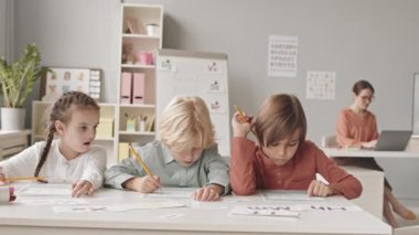 Sınıfta oturmuş konuşma terapisi testleri yapan üç ilkokul öğrencisinin belden aşağısı yavaş çekim yaparken, arka planda dizüstü bilgisayarda çalışan genç bayan öğretmenleri...