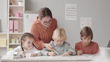 Waist-up slowmo portrait of three cheerful kids of elementary age and young female speech therapist looking at camera while doing exercises on alphabet knowledge in classroom