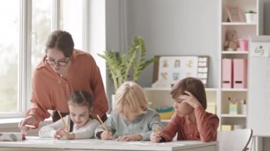 Medium slowmo shot of young female speech therapist helping group of three elementary kids doing exercises on alphabet knowledge, sitting at desk in bright classroom