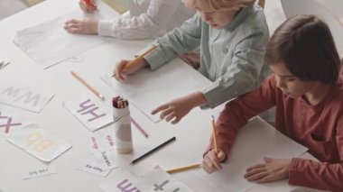 Top view slowmo shot of three elementary children practicing writing skills looking at colorful letter flashcards at desk during speech and language therapy in classroom