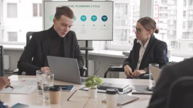 Medium of intelligent Caucasian businesswoman in wheelchair talking to young male colleague, sitting at table in busy conference room at daytime, having financial department meeting