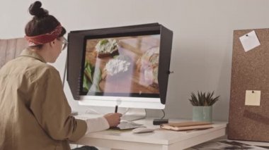 Medium shot of young female food photographer editing photos on computer using graphic tablet and stylus, sitting in her office