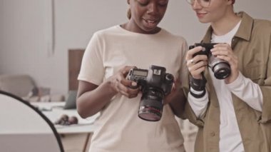 Cropped shot of two multiethnic female photographers with digital cameras in hands looking at display screen discussing pictures, standing in bright photo studio