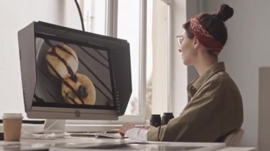 Side-view medium shot of young female photographer editing food picture of delicious cheese pancakes on computer monitor while working in office