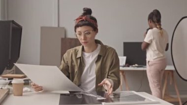 Medium shot of young female photographer looking at printed black and white photos sitting at desk in modern photo studio while her African-American colleague taking pictures in background