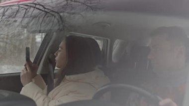 Medium slowmo shot of young Caucasian woman photographing winter forest on smartphone sitting in car with her boyfriend