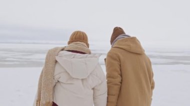 Slowmo shot of young loving couple in winterwear walking on snow on frozen lake holding hands