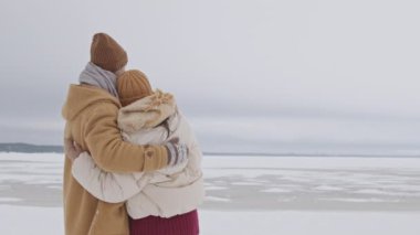 Rear-view medium slowmo shot of young couple hugging each other standing outdoors at snow-covered landscape looking at frozen lake