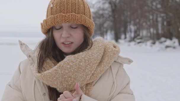 Medium closeup portrait of young Caucasian woman in winterwear looking at camera while warming up her hands standing in snow-covered winter landscape